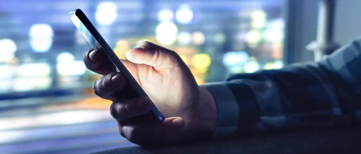 Mão segurando o celular à noite, com as luzes da cidade desfocadas ao fundo